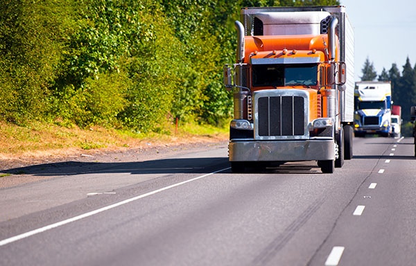 Classic orange semi truck with a refrigerator on a multilane highway road ahead of a convoy of trucks with trailers moving in the right lane highway on a background of green trees and bushes. Movement on the modern highway assures transportation of various goods on these trucks.